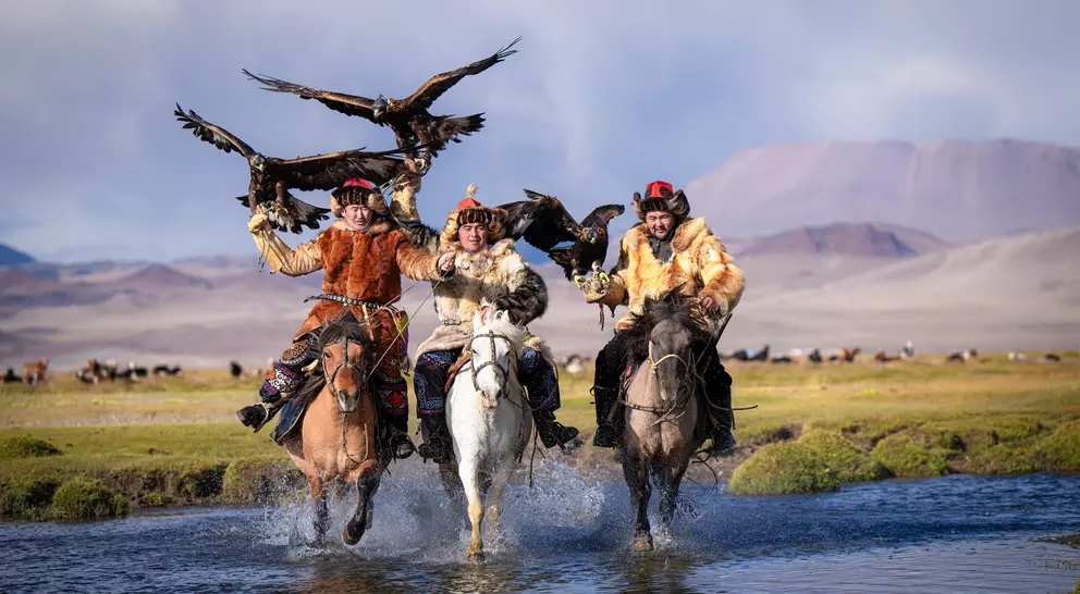 Three riders on horseback splash through a stream, each holding a golden eagle, with mountains in the background.