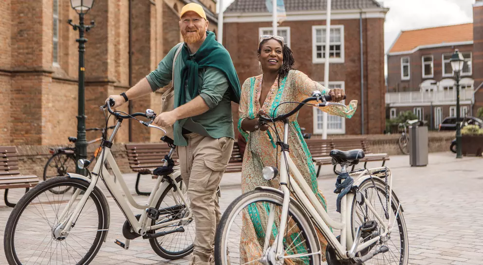 Two smiling people, a man and a woman, stand with bicycles in a charming plaza surrounded by historic buildings.