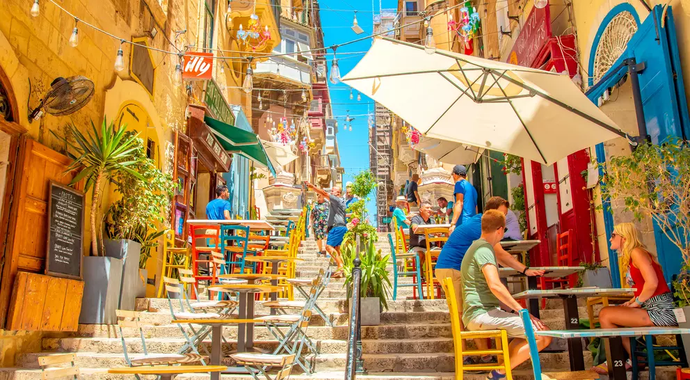 Colorful outdoor cafes line a sunlit, vibrant street, with people enjoying drinks and a bright blue sky overhead.