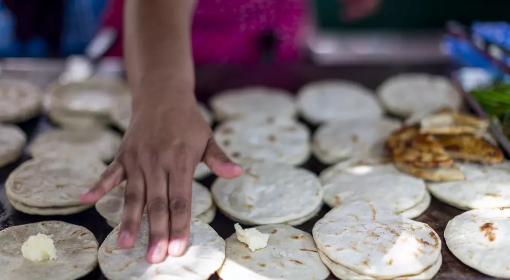 A hand spreads butter on freshly made flatbreads on a cooking surface, surrounded by more flatbreads and some grilled food.