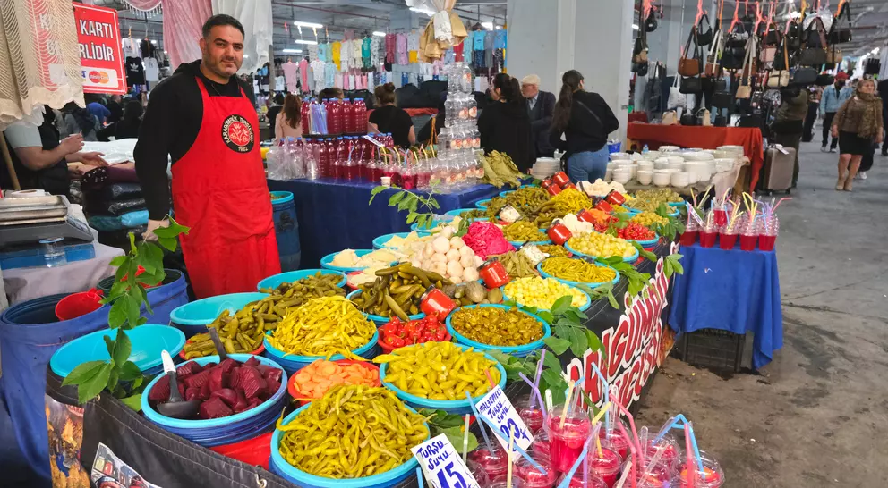 A vendor in an apron stands behind a vibrant table filled with colorful fruits, vegetables, and drinks at a market.