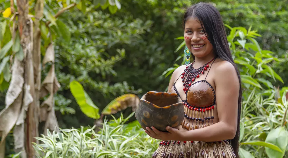 A woman in traditional attire smiles while holding a decorative bowl surrounded by lush greenery.