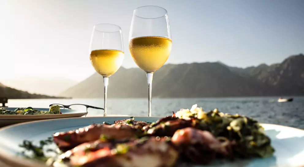 Two glasses of white wine in front of a scenic view with mountains, alongside a plate of seafood and greens.