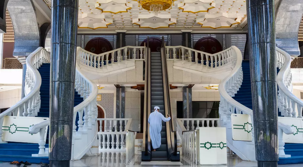 A person in traditional attire stands on an escalator in an ornate hall with grand staircases and a beautifully decorated ceiling.