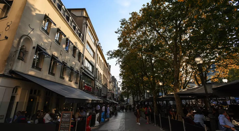 Street lined with cafes and trees, busy with people enjoying dining and conversation in a warm, autumn afternoon.