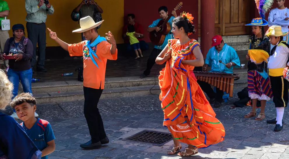Colorful dancers perform a traditional dance in a lively outdoor setting, with musicians and a crowd around them.