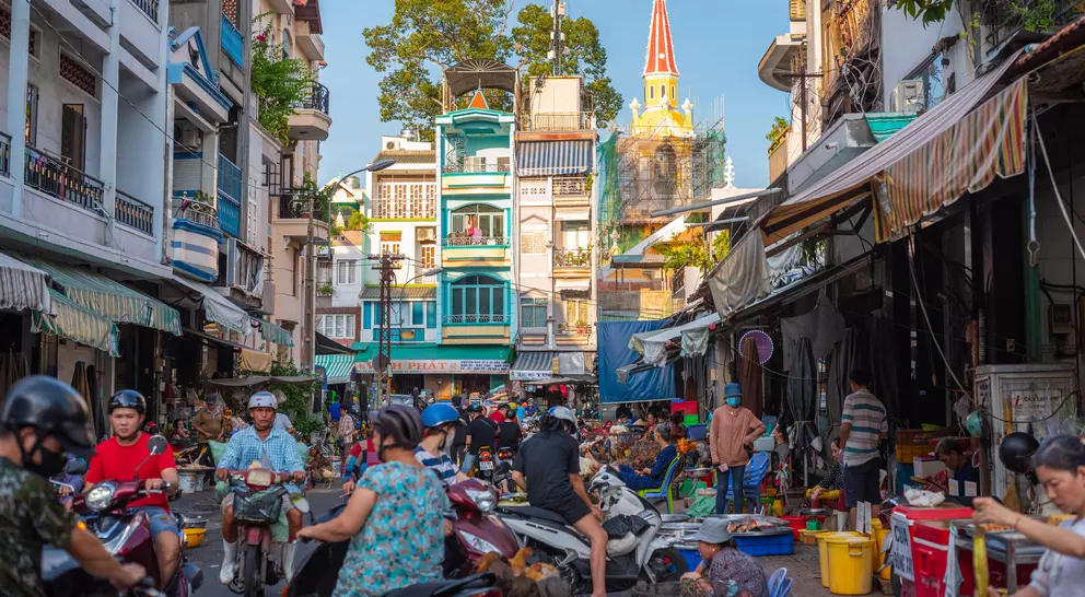 Bustling street scene filled with people and motorbikes, tall buildings, and a church steeple in the background.