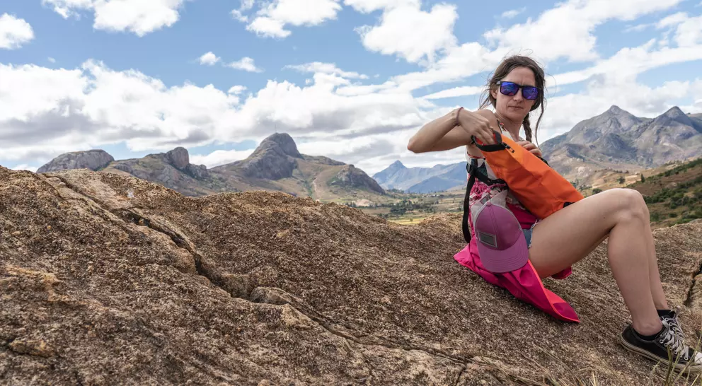 A person wearing sunglasses sits on a rock in the mountains, adjusting their backpack against a scenic backdrop.