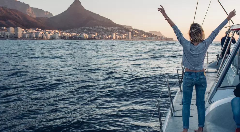 Person on a sailboat raises arms in joy, with mountains and a city skyline in the background at sunset.