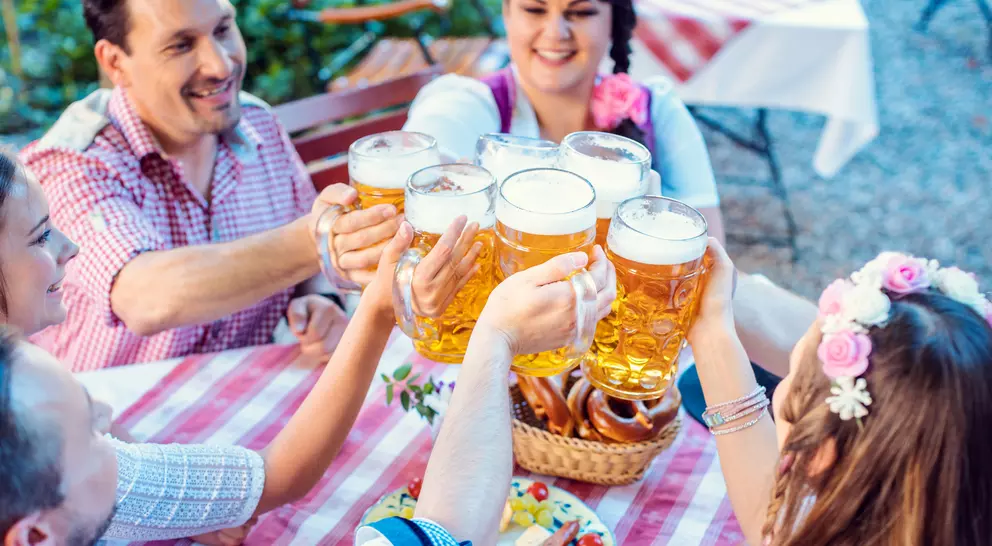 Group of people cheerfully toasting with large beers at a outdoor table, surrounded by pretzels and a festive atmosphere.