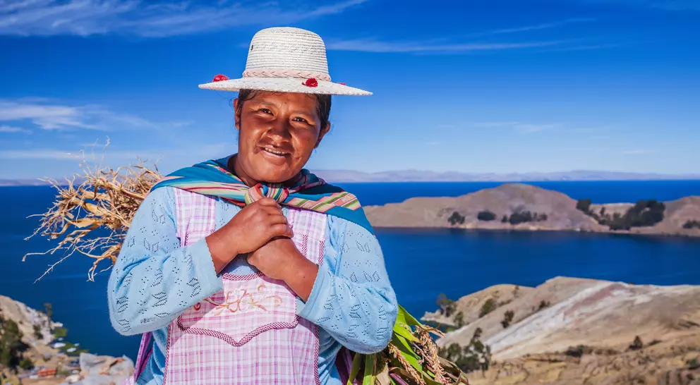A smiling woman in traditional attire stands near a lake, holding plants, with mountains and a clear blue sky in the background.