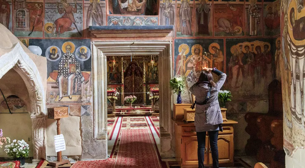 A person admires colorful frescoes inside a church, with floral arrangements and a dimly lit altar in view.
