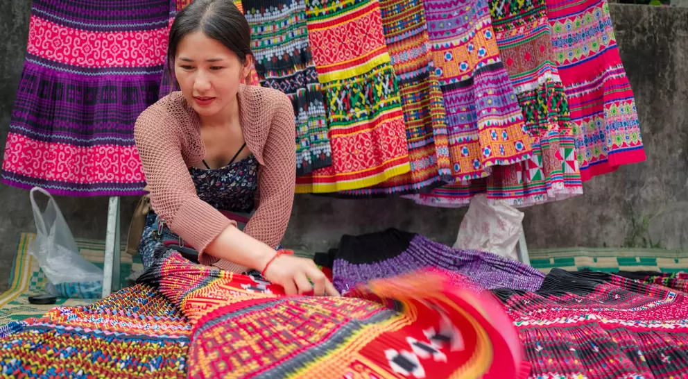 A woman arranges colorful, patterned textiles at a market, with vibrant traditional garments hanging in the background.