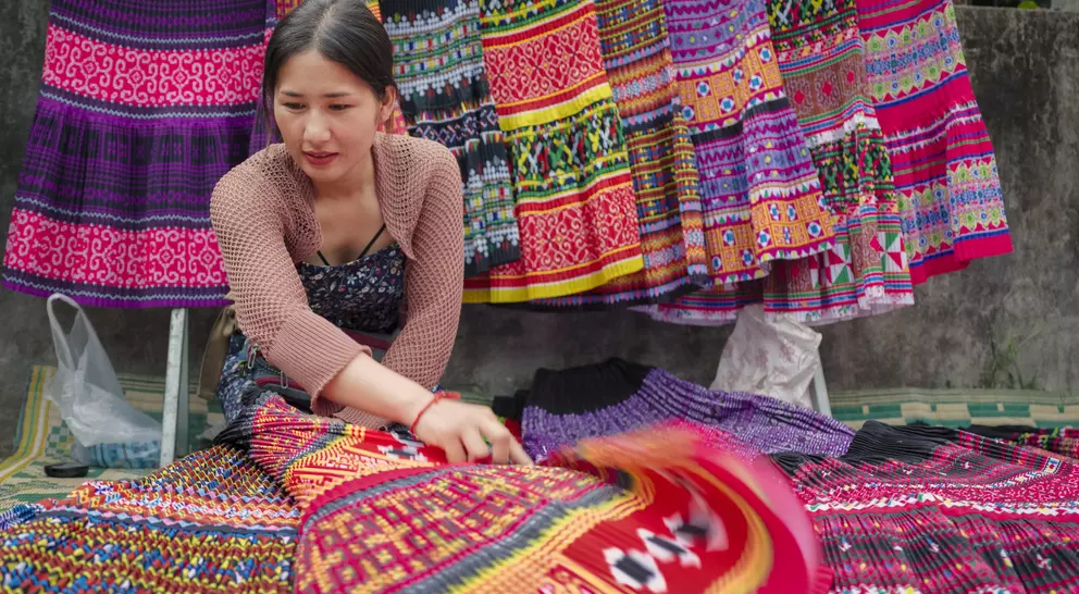 A woman arranges colorful, patterned textiles at a market, with vibrant traditional garments hanging in the background.