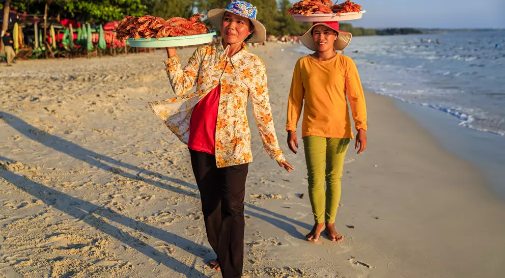 Two women walk along a beach, carrying trays of food on their heads, with a calm sea and trees in the background.