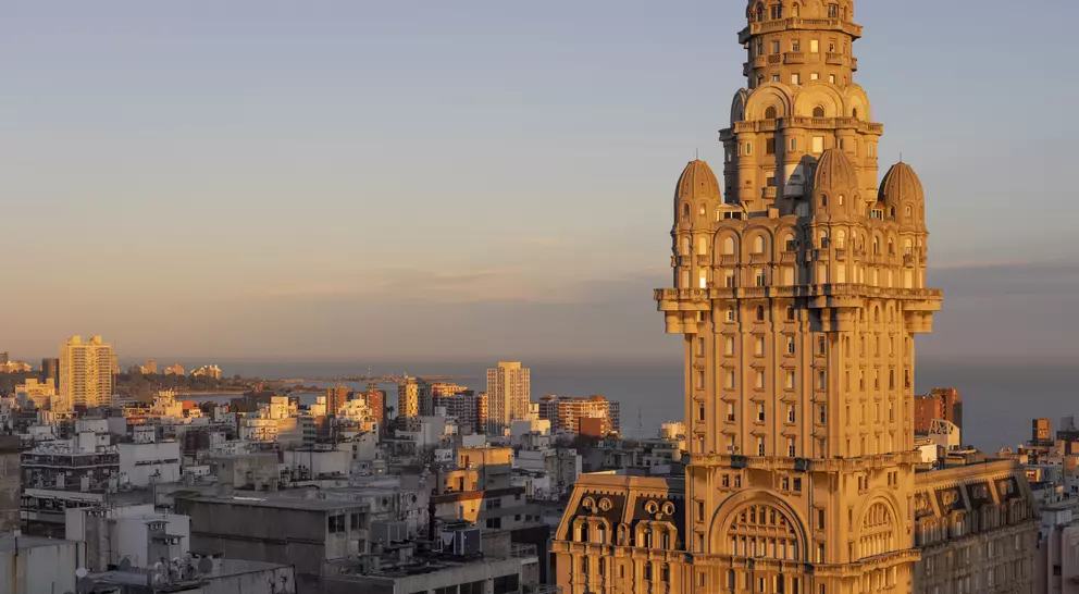 Historic tall building with intricate architecture, bathed in sunset light, overlooking a city skyline and coastline.