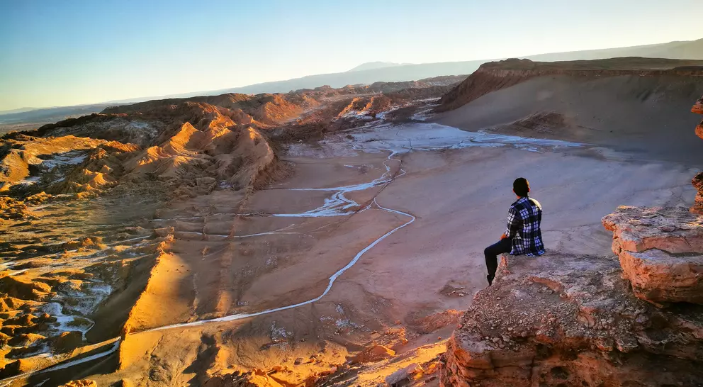 A person sits on a rocky ledge, overlooking a vast desert landscape at sunset.