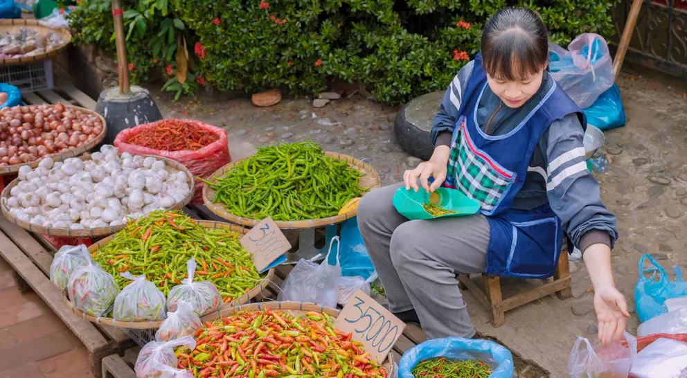 A vendor sits among colorful baskets of vegetables and spices at a market, sorting and preparing fresh produce.