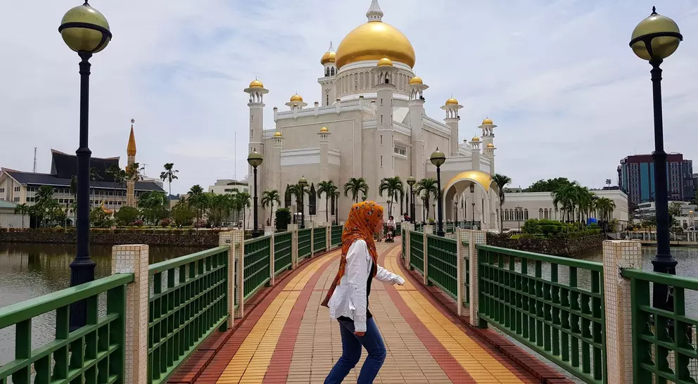 A person with orange hair walks on a bridge toward a grand mosque with golden domes, surrounded by water.