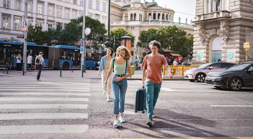 A group of three people walk across a street with a suitcase, surrounded by city buildings and buses.