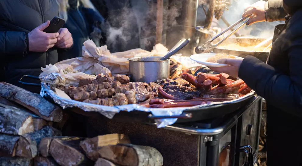 Close-up of a food stall with various meats being served, steam rising, and people in the background.