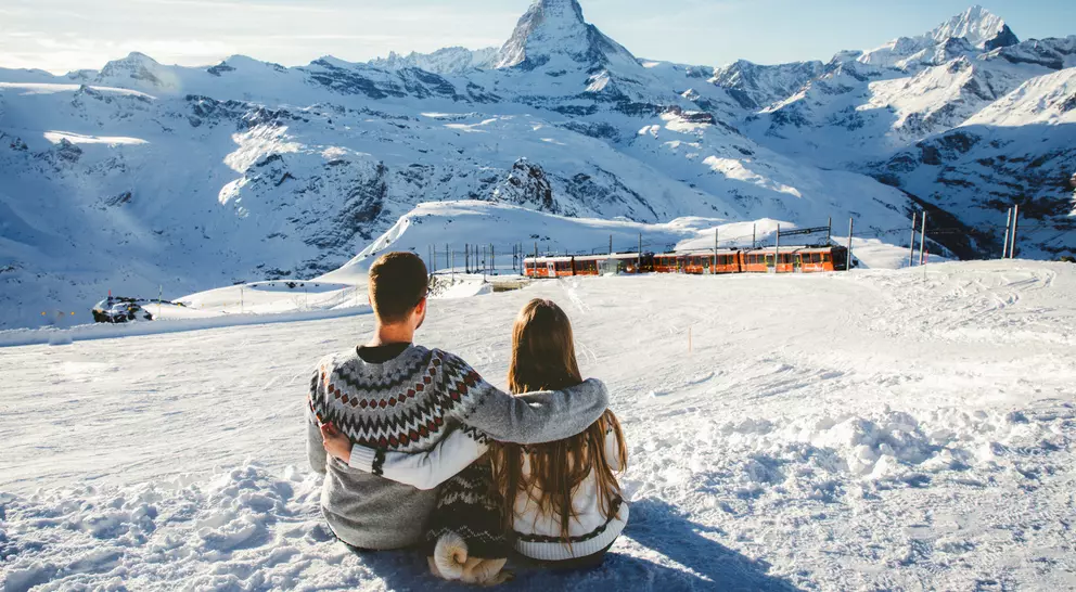 A couple sits together on a snowy slope, overlooking mountains, with the Matterhorn peak in the background.