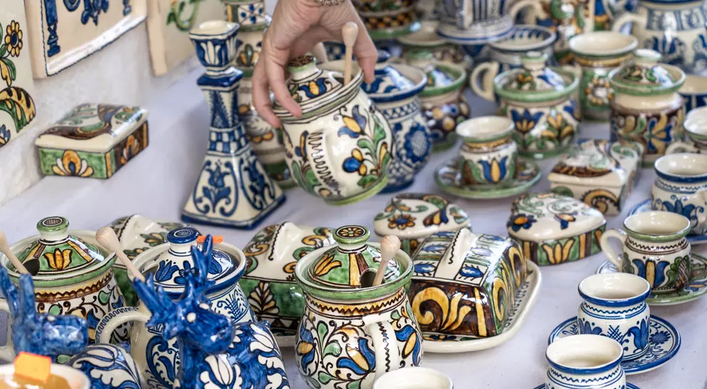 A hand reaches for a colorful, ornate pottery jar among various other decorated ceramics on a display table.