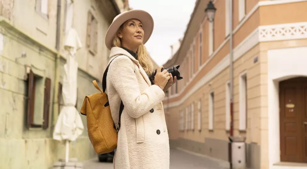 A woman with a camera looks up while standing in a charming, narrow street with colorful buildings.