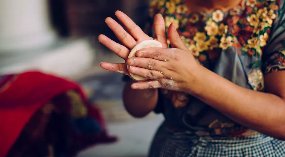 A person is shaping dough in their hands, wearing a colorful floral top with a checkered apron.