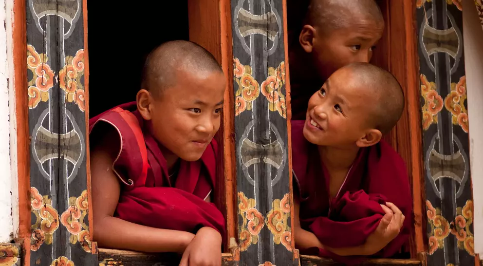 Three young monks with shaved heads peek out from a beautifully decorated window, smiling and engaging with each other.