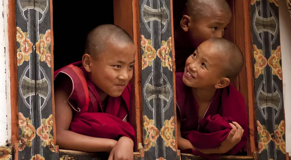 Three young monks with shaved heads peek out from a beautifully decorated window, smiling and engaging with each other.