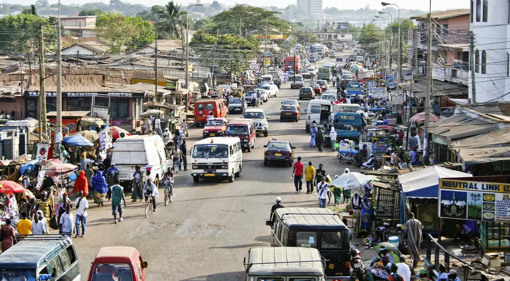 Busy street scene in a town, filled with vehicles, vendors, and pedestrians amidst shops and buildings.