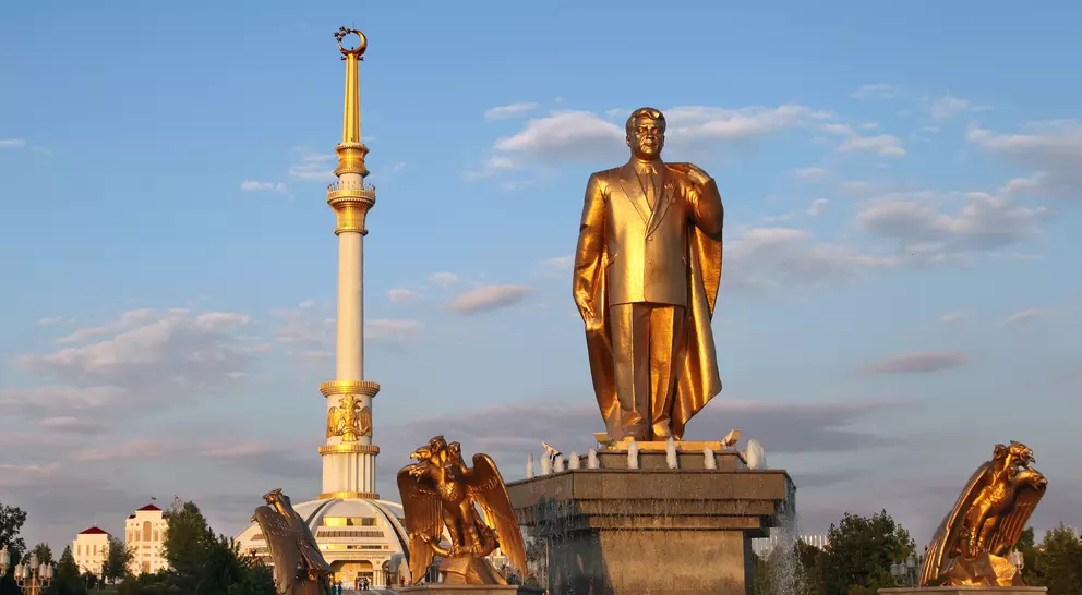 Golden statue of a man with a cape, surrounded by fountains, and a tall spire in the background against a blue sky.