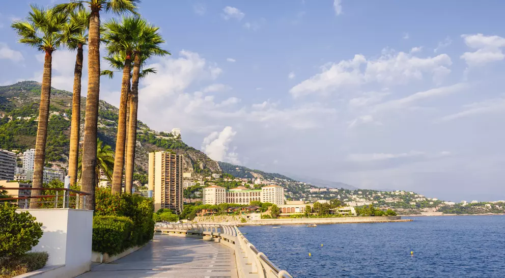 Scenic view of a coastal promenade lined with palm trees, buildings, and mountains under a blue sky with clouds.