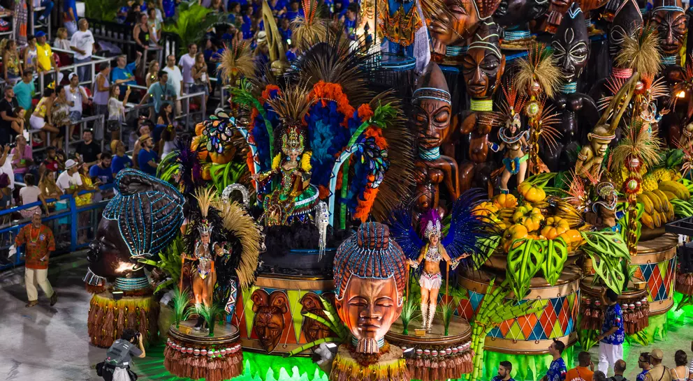 Colorful parade float featuring intricate masks and tropical motifs, surrounded by a crowd in vibrant attire.