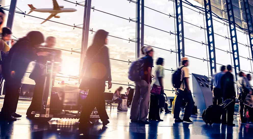 Blurred travelers moving through a bustling airport terminal, with an airplane flying outside the large glass windows.