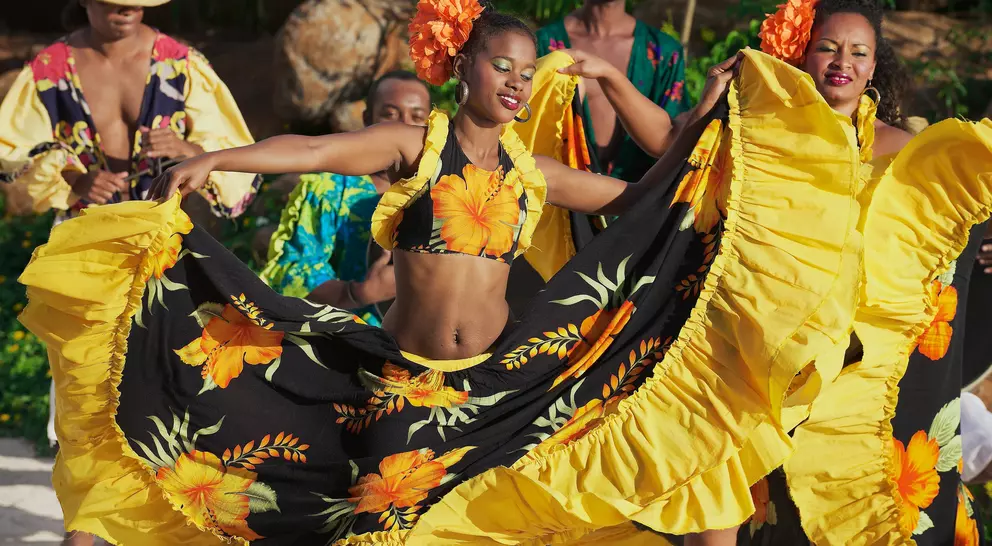 Two women in vibrant yellow and black dresses dance joyfully, surrounded by musicians dressed in colorful attire.