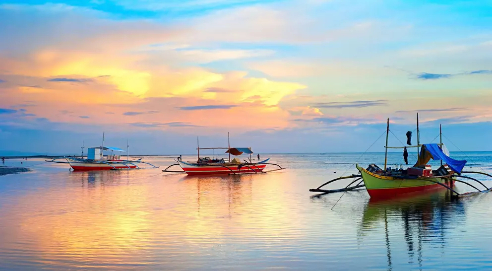 Three colorful boats on calm water under a vibrant sunset sky with clouds reflecting in the water.