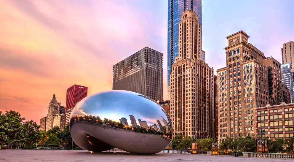 Cloud Gate sculpture reflects the skyline at sunset, surrounded by urban buildings in a vibrant city landscape.