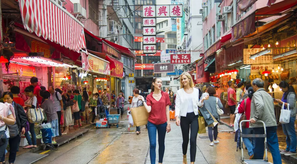 Two women walk through a bustling market street filled with colorful shops and people, vibrant signs in the background.