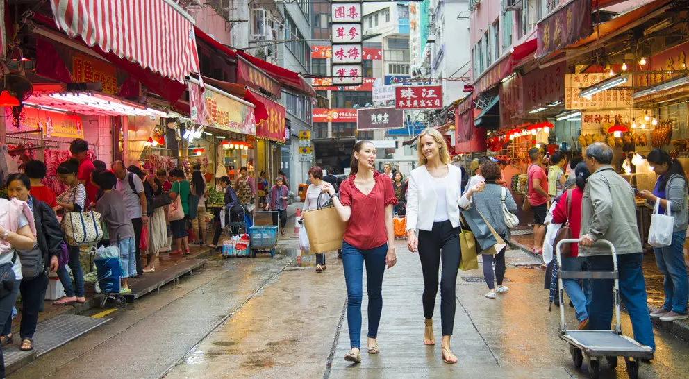 Two women walk through a bustling market street filled with colorful shops and people, vibrant signs in the background.