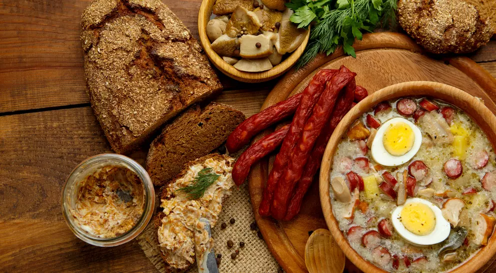 A rustic wooden table displays a hearty meal with soup, sliced sausage, bread, pickles, and garnishes.