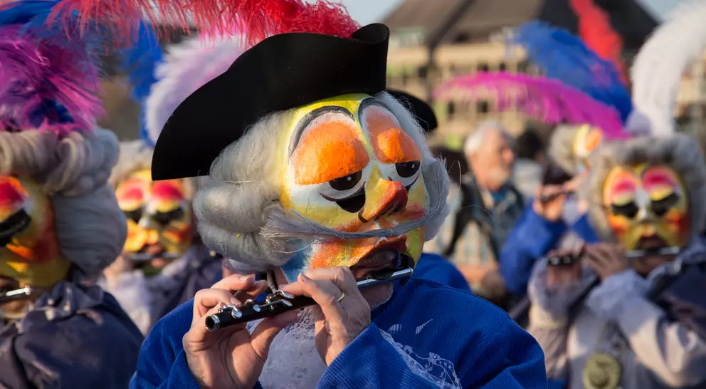 A performer in a feathered hat plays a flute, surrounded by others in colorful masks and costumes during a celebration.