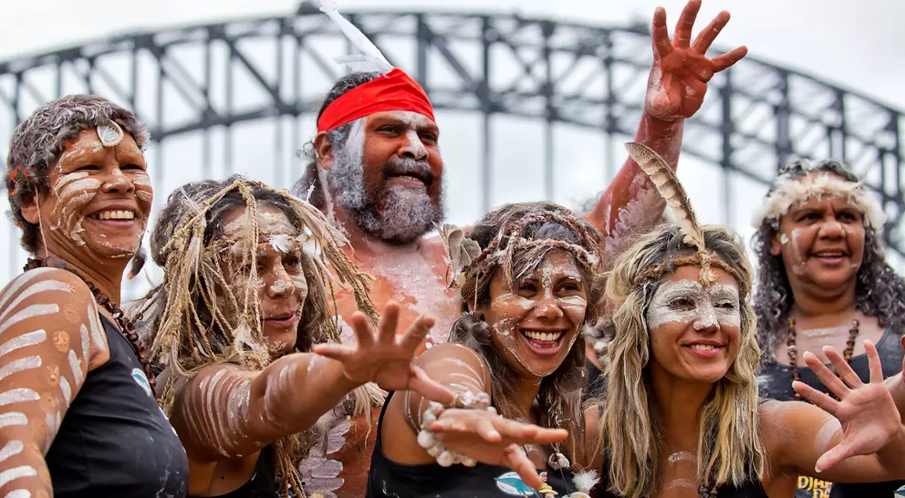 A group of six Indigenous people celebrating with traditional attire and body paint in front of a bridge.
