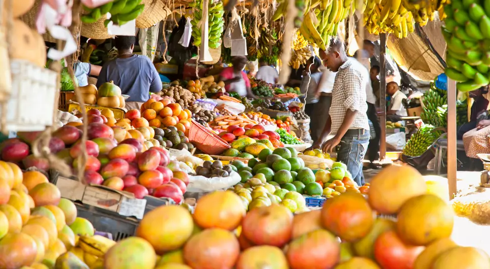 A vibrant market stall filled with various fruits and vegetables, with shoppers browsing under a canopy of greenery.