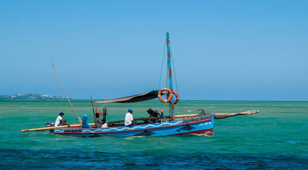A colorful fishing boat with several people on the water, surrounded by a bright blue sky and clear green sea.
