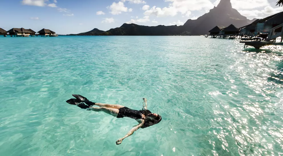 Person in snorkel gear floats peacefully in turquoise water, surrounded by mountains and overwater bungalows under a sunny sky.