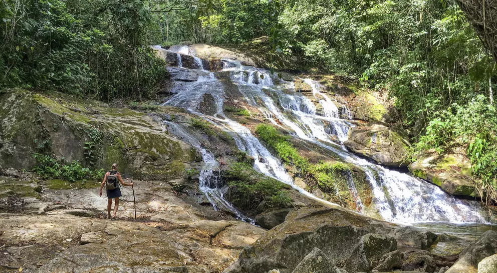 A hiker stands on rocky terrain near a cascading waterfall surrounded by lush greenery.