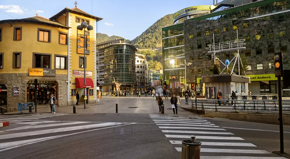 A city square with a mix of modern and traditional buildings, pedestrians crossing a striped crosswalk under a clear sky.