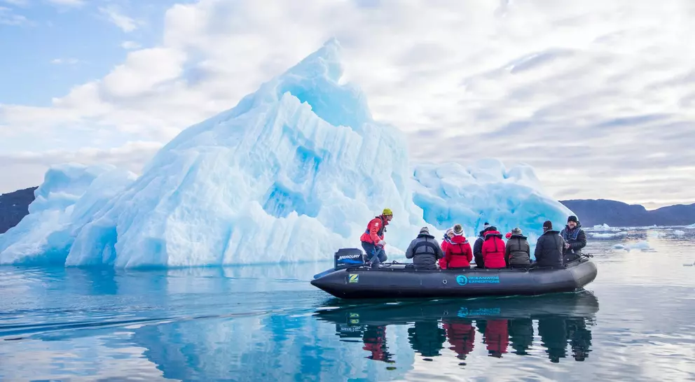 A group of people in a boat near a large blue iceberg on calm water under a cloudy sky.